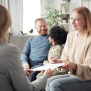 Happy young woman signing contract about adoption while talking to social worker with her family in background
